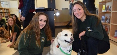 Two female St.Benedict students stand smiling with a therapy dog.