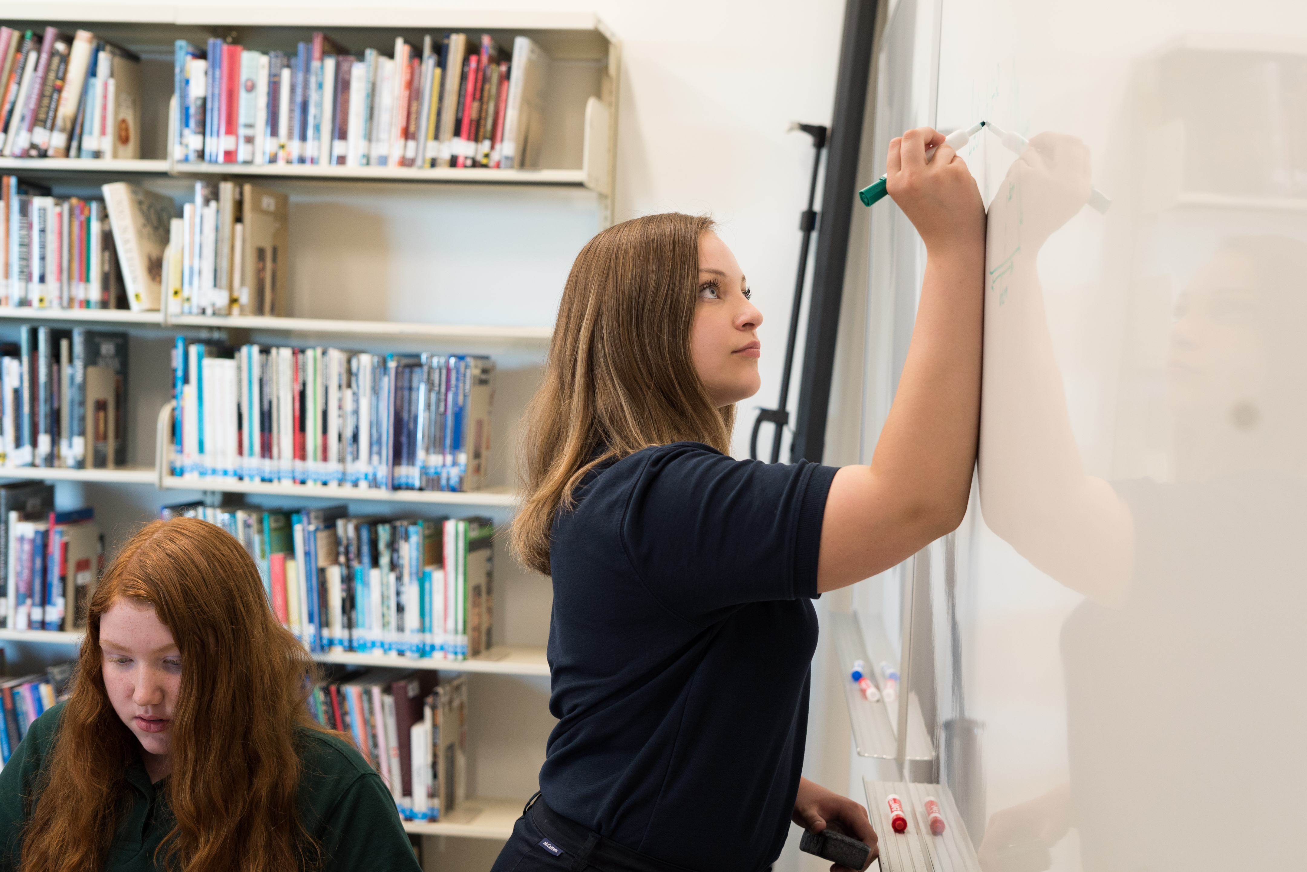 A girl draws a math problem on a white dry erase board as another student watches.